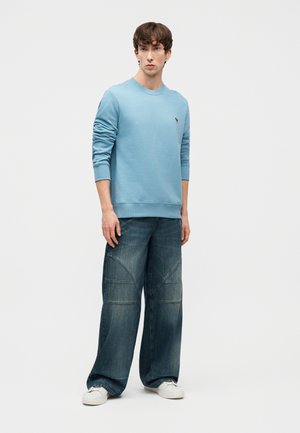 Noor mees seisab, kandes helesinist dressipluusi, laia säärega tuhmiks kulunud teksaseid õmblustega ja valgeid tossusid, taust on lihtne.