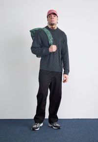 Pulover gri cu model tricotat, pantaloni negri și teniși gri-negru-verzi. Rucsac verde pe umăr, șapcă roz. Fundal neutru.