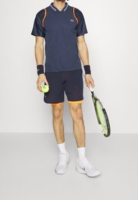 Marinblå kortärmad tennis-polo med orange detaljer, matchad med marinblå shorts med en orange fåll, vita tennisskor, håller en racket och en boll.