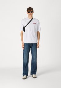 Camiseta blanca de manga corta con gráfico rojo, jeans de mezclilla azul, bolso cruzado negro, zapatos grises y blancos, y gafas de sol negras.
