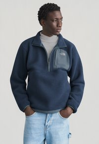 Άνδρας που φοράει μπλε σκούρο φλις με φερμουάρ στο τσέπη στο στήθος και ανοιχτόχρωμο τζιν, στέκεται με τα χέρια στις τσέπες, κοιτάζοντας πλάγια.