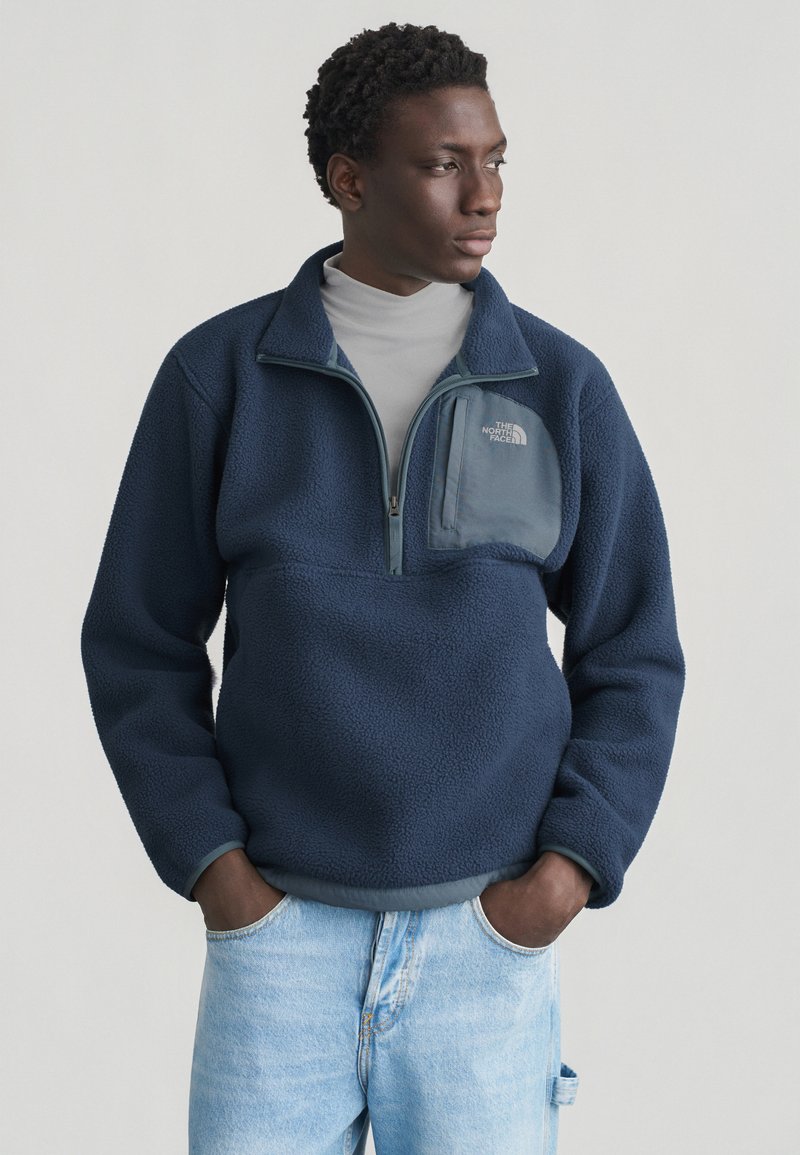 Άνδρας που φοράει μπλε σκούρο φλις με φερμουάρ στο τσέπη στο στήθος και ανοιχτόχρωμο τζιν, στέκεται με τα χέρια στις τσέπες, κοιτάζοντας πλάγια.