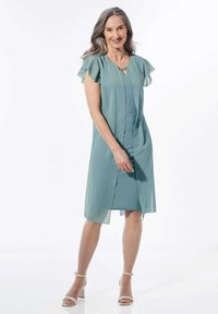 Ein türkisfarbenes, knielanges Kleid aus leichtem Stoff mit schwingenden Ärmeln und einer teilweise geknöpften Vorderseite, das ein kleines Schlüsselloch-Detail am Hals aufweist.