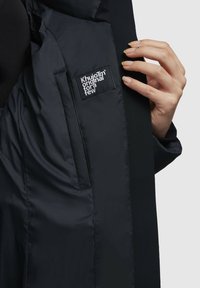 Čierny zateplený kabát má hladké, saténové podšívkové tkaniny s viditeľným štítkom. Ruka držiaca podšívku odhaľuje elegantný dizajn a textúru materiálu.