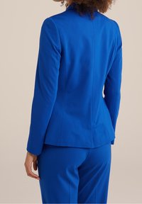 Blazer aderente di un blu reale con una texture liscia, spalle strutturate e una delicata cucitura in vita. Presenta maniche lunghe e un classico colletto a punta.