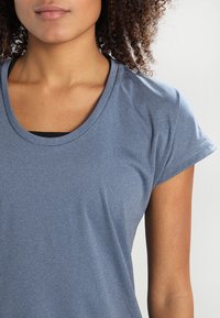 Modré sportovní tričko s kulatým výstřihem, krátkými rukávy a hladkým povrchem, částečně viditelné na světlém pozadí.