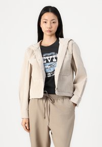 Veste en velours côtelé beige avec capuche, boutons-pression et deux poches avant, portée par-dessus un t-shirt graphique noir et un pantalon beige à cordons.