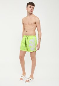 Pantaloncini da nuoto verde neon con un grande motivo floreale bianco, vita con cordoncino e tessuto leggero. Indossati con sandali bianchi a scivolo.