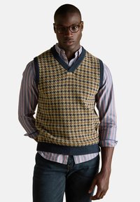 Kootud vest V-kaelusega halli ja meresinise šakkmustriga, kantud triibulise sinise ja valge särgi peal, koos tumedate teksadega.