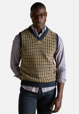 Knitted vest with a v-neck in a houndstooth pattern of tan and navy, worn over a striped blue and white shirt, paired with dark jeans.