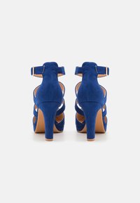 Blue suede high-heeled shoes with a back ankle strap and crisscrossed front design. The heel is approximately 4 inches tall.