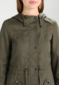 Olivgrön kapuschongjacka med tryckknappar, dragsko i midjan och fickor med dragkedja i ett texturerat tyg. Avslappnad och funktionell design.