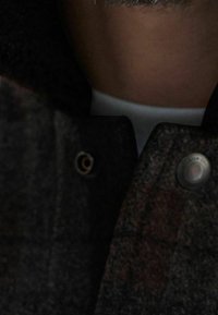 Czarny szary wełniany płaszcz o teksturowanym wykończeniu, z srebrnym guzikiem i dużym dziurkiem na guzik. Kołnierz jest lekko złożony w dół.