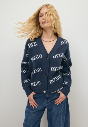 Vrouw met golvend blond haar draagt een marineblauwe cardigan met witte "BIZUU" tekst en blauwe spijkerbroek, staand tegen een eenvoudige lichte achtergrond.