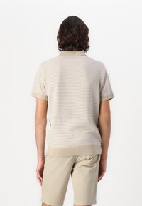 Beige stripete polo skjorte med korte ermer og ribbestrikket krage, som viser en myk tekstur og en tettsittende design, kombinert med lyse shorts.