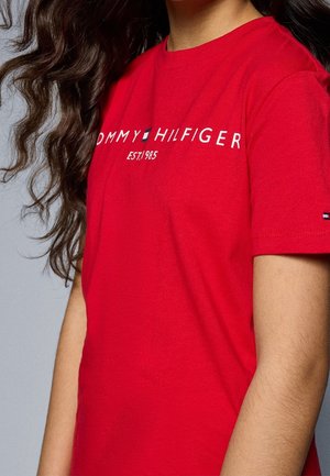 Person iført en klar rød Tommy Hilfiger t-shirt med hvid logo tekst og et lille flag på ærmet, med langt mørkt bølget hår synligt.