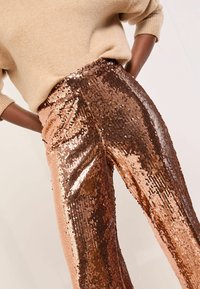 Személy fényes réz színű flitteres nadrágban és bézs pulóverben, kezei a csípőjén pihennek, egy világos, egyszínű háttér előtt.