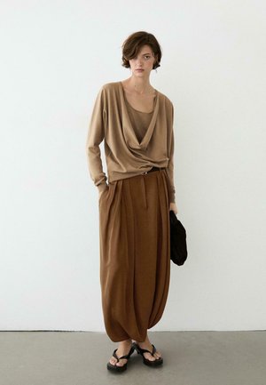 Jonge vrouw die tegen een witte muur staat, gekleed in een losvallende bruine gedrapeerde top, bijpassende brede geplooide broek, zwarte sandalen met hakken en een zwarte clutch vasthoudend.