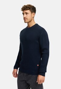 Granatowy sweter dzianinowy o prążkowanej strukturze, okrągłym dekolcie i długich rękawach. Zawiera małą naszywkę z logo na dolnym lewym rogu.