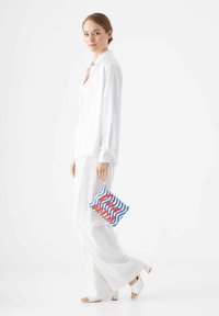 Witte blouse, witte broek en witte hoge hakken. Draagt een kleurrijke clutch met een blauw en rood golvend ontwerp en tekst.