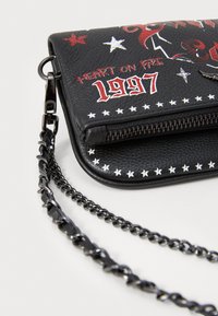 Sac en simili cuir noir avec des motifs d'étoiles blanches et des graphismes rouges, doté d'une fermeture éclair et d'une bandoulière en chaîne noire. Accentué avec "Cœur en Feu" et "1997".