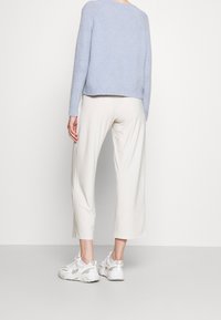 Žena v světle modrém svetru, krémových širokých zkrácených kalhotách a bílých robustních teniskách stojí proti čisté bílé zdi.