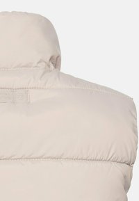 Veste beige matelassée vue de dos montrant un col montant et des détails de coutures horizontales sur un tissu lisse.