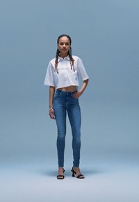 Chemise blanche courte à manches courtes, associée à un jean bleu ajusté. Le mannequin porte des sandales à talons noires et a les cheveux tressés. Fond uni.