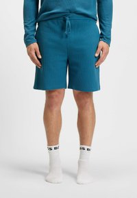 Teal shorts van een textuurstof met een elastische tailleband met trekkoord. Draagt witte sokken met zwart logo-tekst.