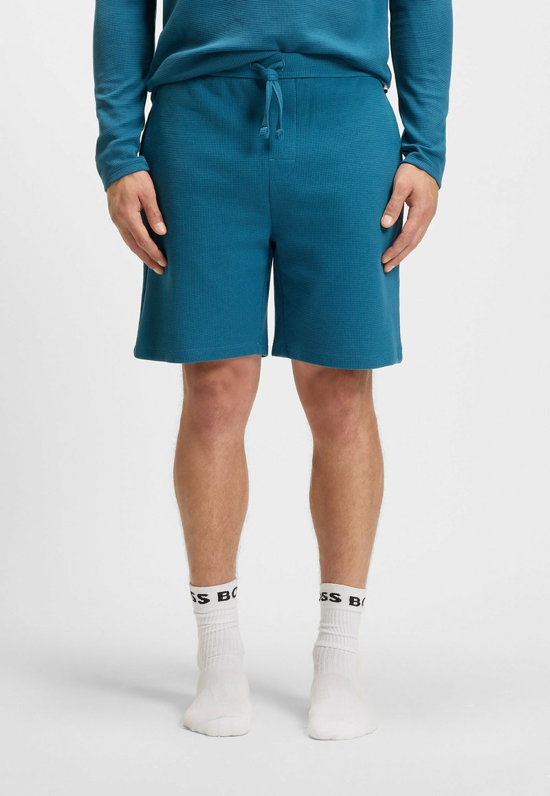Teal shorts van een textuurstof met een elastische tailleband met trekkoord. Draagt witte sokken met zwart logo-tekst.