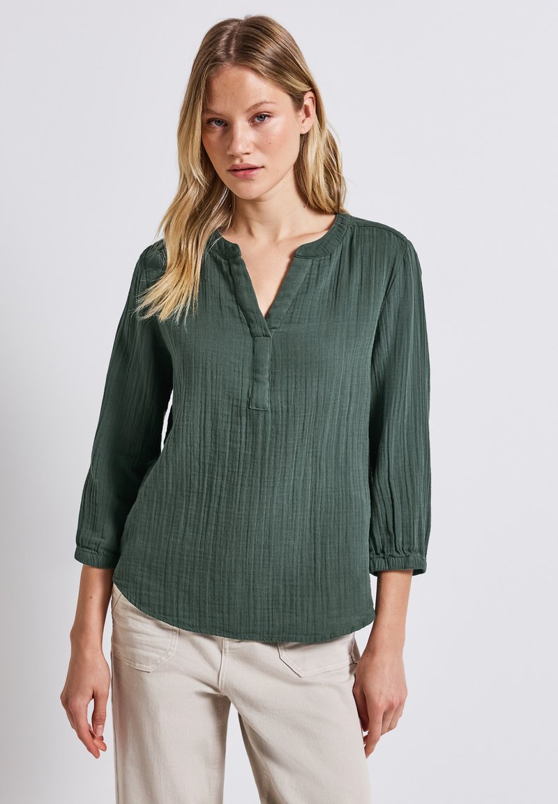 Blusa verde, con texture, a scollo a V, maniche a tre quarti e vestibilità morbida. Presenta sottili pieghe e un orlo arrotondato.