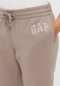 Gai brūni sporta bikses, kas izgatavotas no mīksta kokvilnas, ar elastīgu jostu un auklu, uz kuras priekšpuses ir izšūts "GAP" gaiši rozā krāsā.