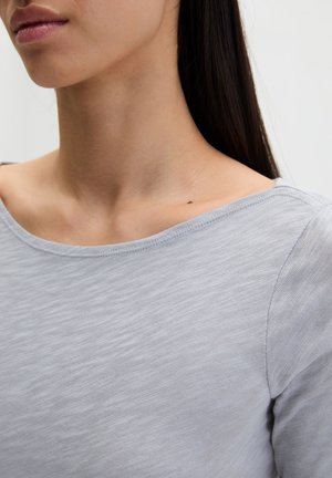 Zbliżenie na szyję i ramię osoby ubranej w jasnoszary, teksturowany top z długim rękawem i szerokim dekoltem.