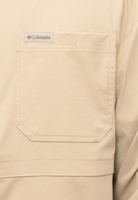 Τσέπη πουκαμίσου Columbia σε μπεζ χρώμα με εμφανείς ραφές και μια μικρή υφασμάτινη ετικέτα με το λογότυπο της εταιρείας ραμμένη πάνω από το άνοιγμα της τσέπης.