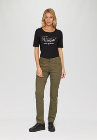 Zwarte korte mouwen shirt met de opdruk "JE POTENTieel kent GEEN grenzen", gecombineerd met olijfgroene slim-fit broek en zwarte sneakers.