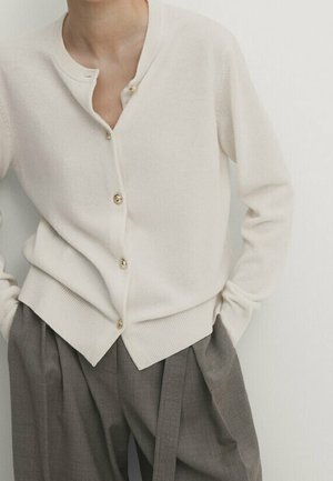 Vrouw met een crèmekleurig vest met knopen en geribbelde randen, gecombineerd met loszittende grijze broek met hoge taille en een stoffen riem.