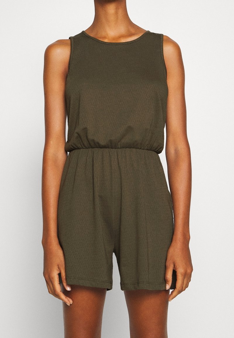 Combinaison sans manches vert olive avec taille élastique, portée par une personne debout, les bras détendus le long du corps, sur un fond uni.
