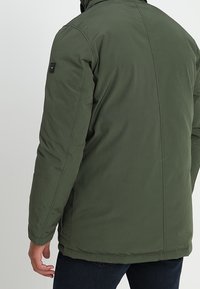 Veste isolante verte avec un col haut, présentant un patch logo sur l'épaule et une texture lisse et mate.