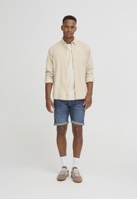 Chemise beige à boutons sur un T-shirt de couleur claire, associée à un short en jean retroussé. Baskets marron avec lacets blancs et chaussettes mi-mollet.