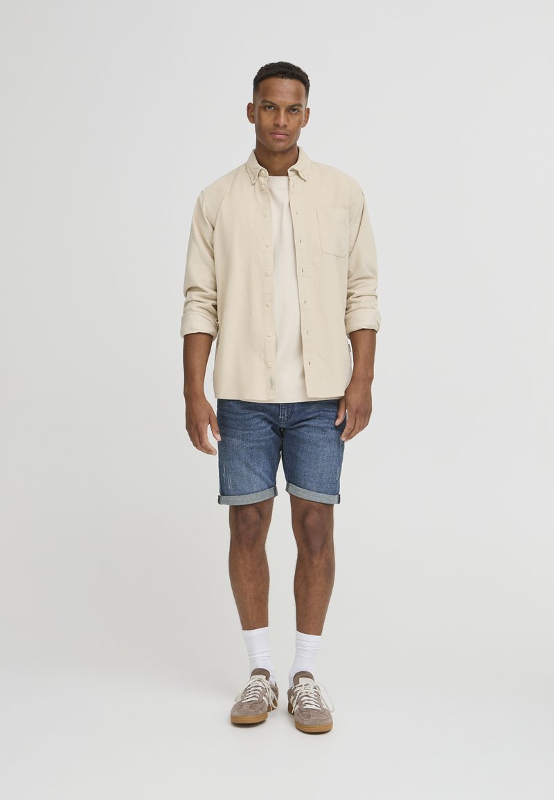 Chemise beige à boutons sur un T-shirt de couleur claire, associée à un short en jean retroussé. Baskets marron avec lacets blancs et chaussettes mi-mollet.