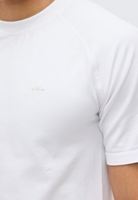 Κοντινό πλάνο ενός ατόμου που φοράει απλό λευκό αθλητικό κοντομάνικο μπλουζάκι με ένα μικρό λευκό λογότυπο στο στήθος.