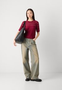 Weinrotes, gestricktes Oberteil mit Ausschnitten, kombiniert mit hellblauen, weit geschnittenen Jeans. Das Modell hält eine schwarze Ledertasche und trägt schwarze Schuhe.