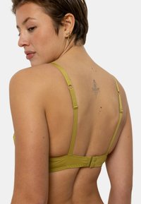 Soutien-gorge vert olive en tissu doux, avec bretelles réglables et fermeture à crochets. Le dos présente un design de tatouage au-dessus de l'épaule gauche.