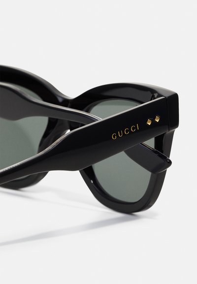 Melni saulesbrilles ar izteiksmīgu kaķa acs formu, zaļiem stikliem un gludu, spīdīgu apdari. Zelta "GUCCI" logo uz kājiņas ar akcentētām detaļām.