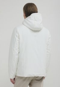 Weiße Kapuzenjacke mit glatter, gepolsterter Textur. Sie verfügt über eine lockere Passform, nahtlose Schultern und subtile Steppdetails.