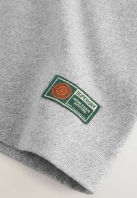 Graues Sweatshirt-Gewebe mit glatter Textur und einem sichtbaren grünen und orangefarbenen Etikett, auf dem "Superdry Vintage Athletic Outfitters" steht.