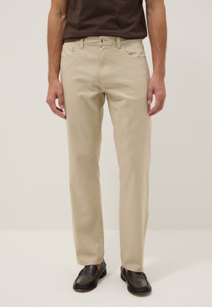 Uomo che indossa pantaloni beige a gamba dritta con tasche frontali e mocassini neri su sfondo semplice.