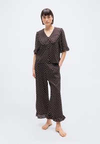 Femme debout, pieds nus, portant un pyjama marron foncé à pois avec un large col et des manches boutonnées à volants ainsi que des ourlets de pantalon volantés.