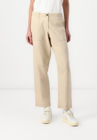 Pantaloni in cotone beige con gamba dritta, tasche frontali e chiusura con bottone, abbinati a sneakers bianche. Design minimale e texture liscia.