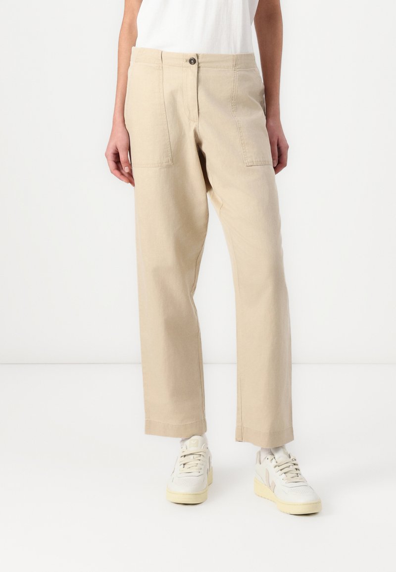 Pantaloni in cotone beige con gamba dritta, tasche frontali e chiusura con bottone, abbinati a sneakers bianche. Design minimale e texture liscia.